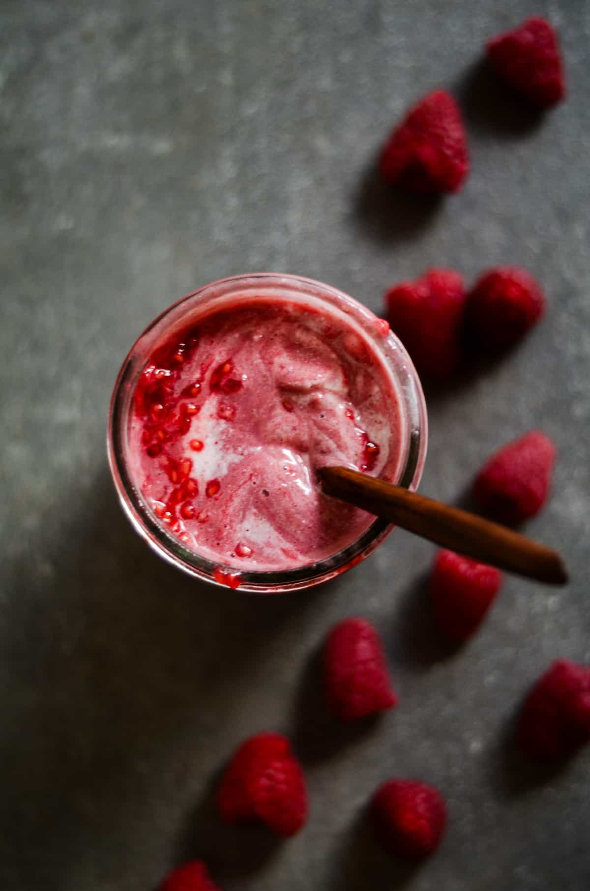 A bright pink raspberry smoothie in a jar with a spoon in it and raspberries beside it.