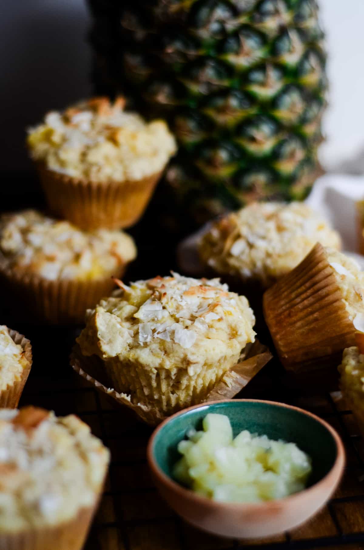 Yellow muffins with a little bowl of crushed pineapple and a whole pineapple behind the muffins.