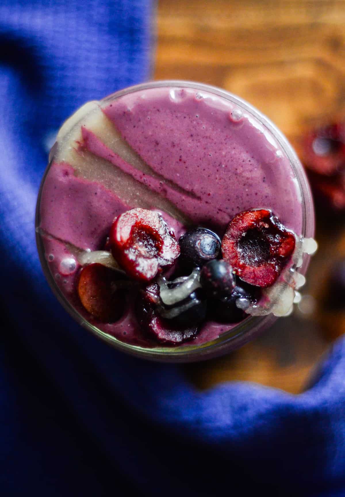 A purple smoothie in a glass with honey, halved cherries, and blueberries on top.