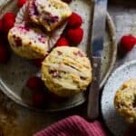 Raspberry muffin on a plate with a knife and raspberries beside it.