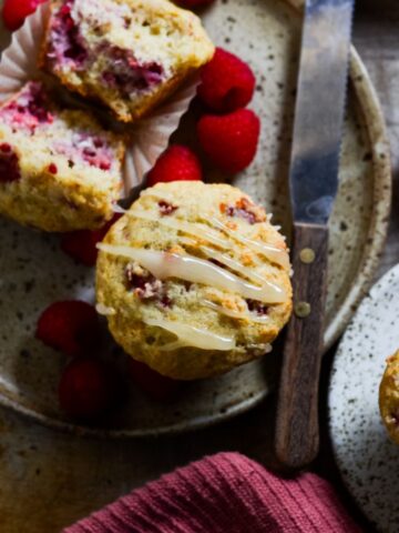 Raspberry muffin on a plate with a knife and raspberries beside it.