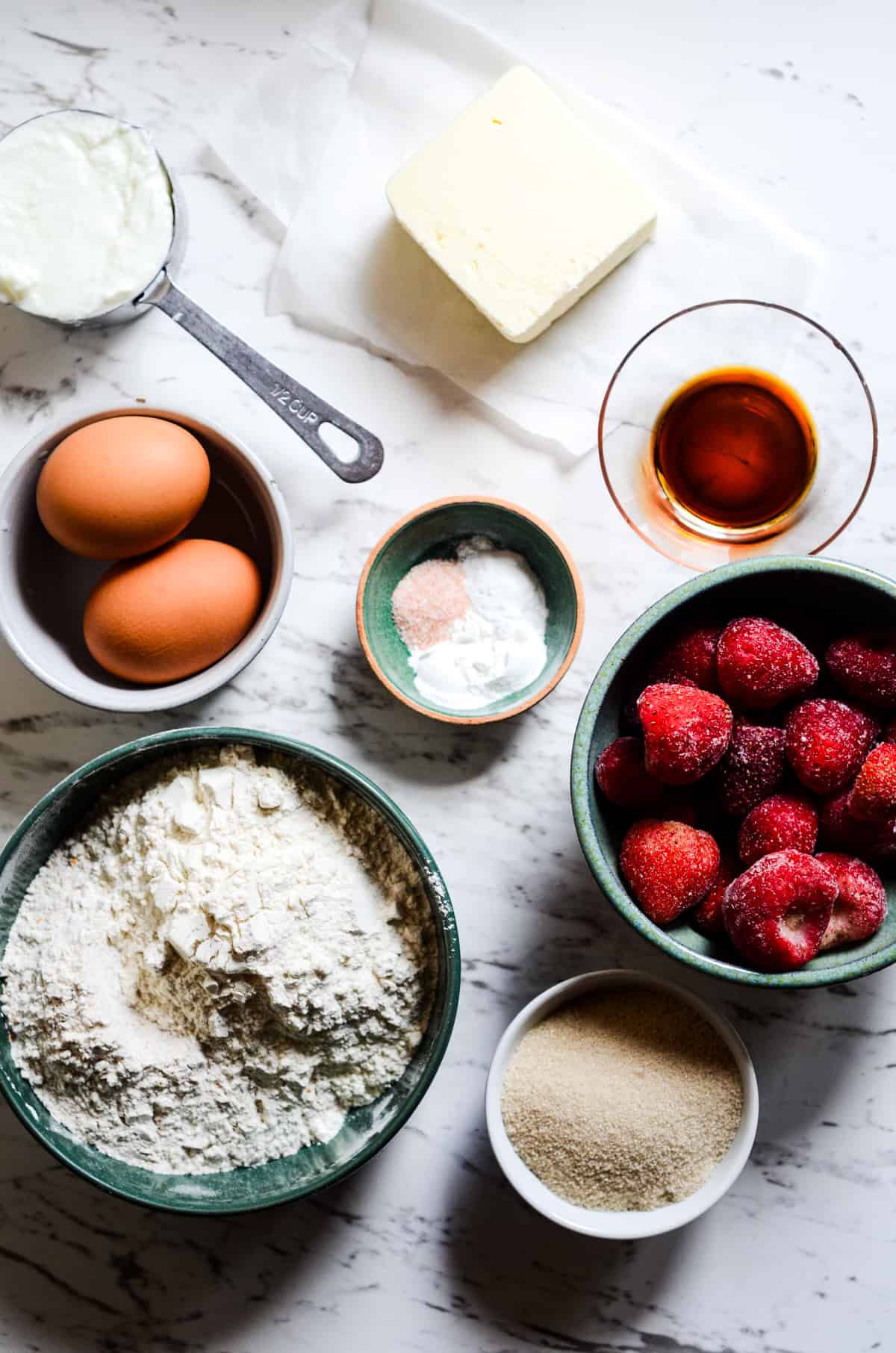 Ingredients for muffins in bowls including flour, eggs, yogurt, butter, salt, baking soda, baking powder, vanilla, cane sugar, frozen strawberries.