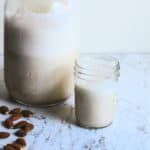 Almond milk in a small glass with a jar of almond milk and almonds beside it.