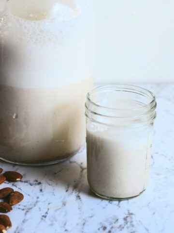 Almond milk in a small glass with a jar of almond milk and almonds beside it.