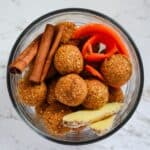 Orange brown carrot cake energy balls with ginger root, fresh carrot, and cinnamon sticks in a glass bowl.