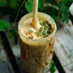 A glass cup filled with light brown smoothie with pumpkin seeds on top, with leaves in the backgrounds.