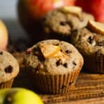 A light brown muffin with mini chocolate chips and apple chunks on top, sitting on a cutting board with apples and muffins beside it.