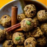 Light brown energy bites with flecks of oats and dark chocolate chips with pumpkin seed on top and cinnamon sticks beside it.