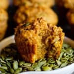 A pumpkin colored muffin with pumpkin seeds on top and sitting in a dish of olive green pumpkin seeds.