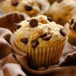 A light colored muffins with a lot of chocolate chips poking out of it, sitting on a brown napkin, with other chocolate chip muffins around it.