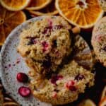 A light orange muffins with red cranberries poking through on a little plate with orange slices behind it.