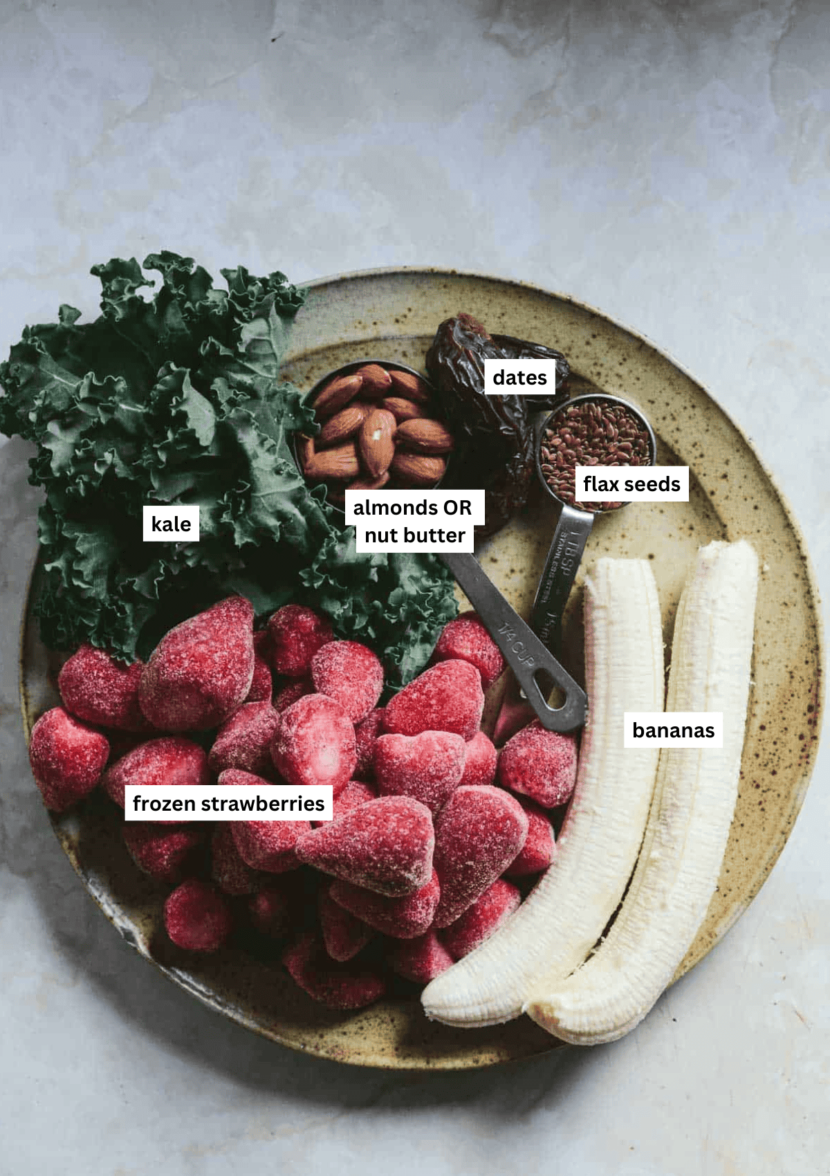 The ingredients needed for a strawberry banana smoothie on a plate, including kale, dates, almonds, flax sees, frozen strawberries, and fresh bananas.