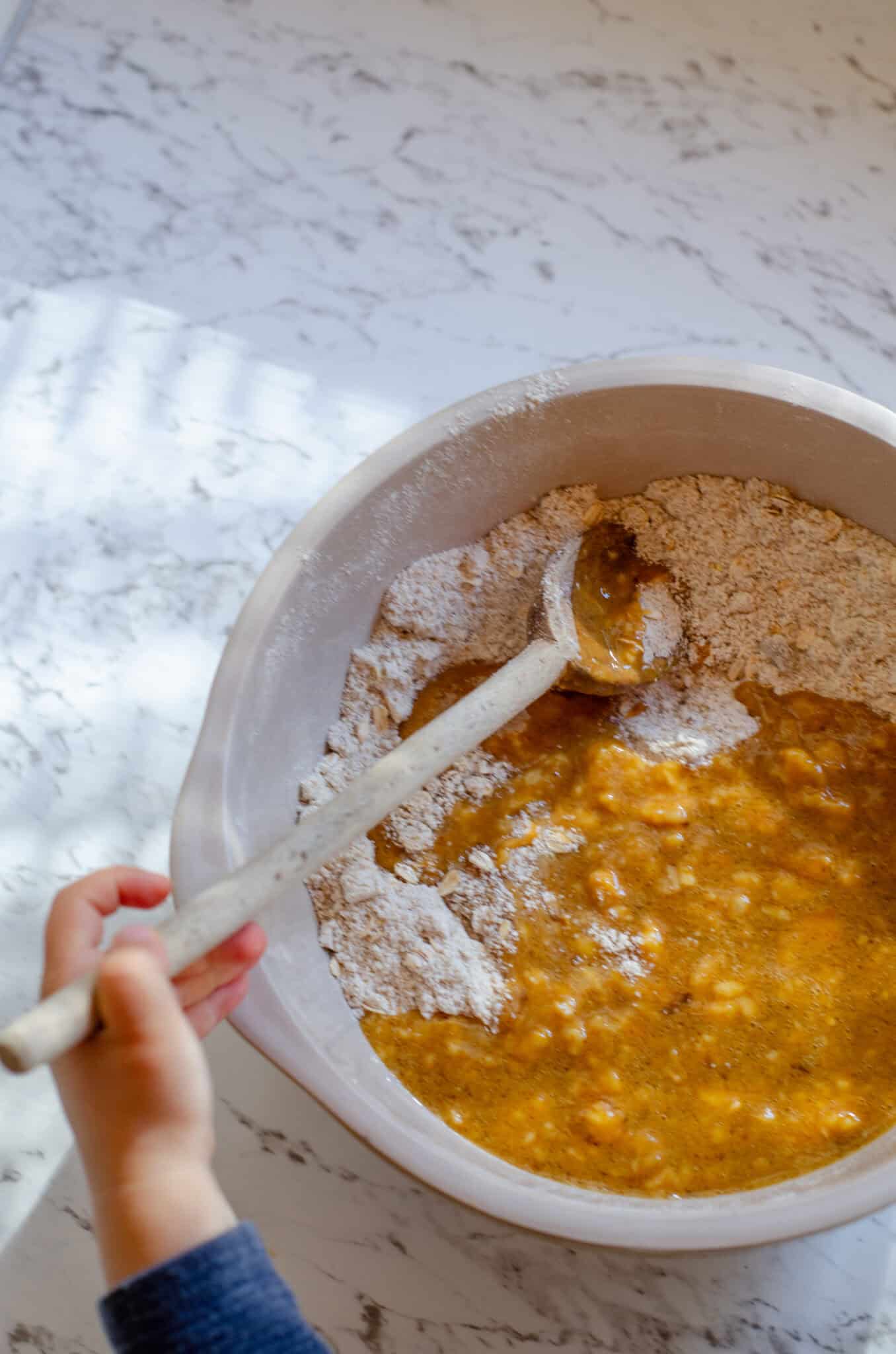 A brown wet mixture being mixed into the flour mixture with a wooden spoon.