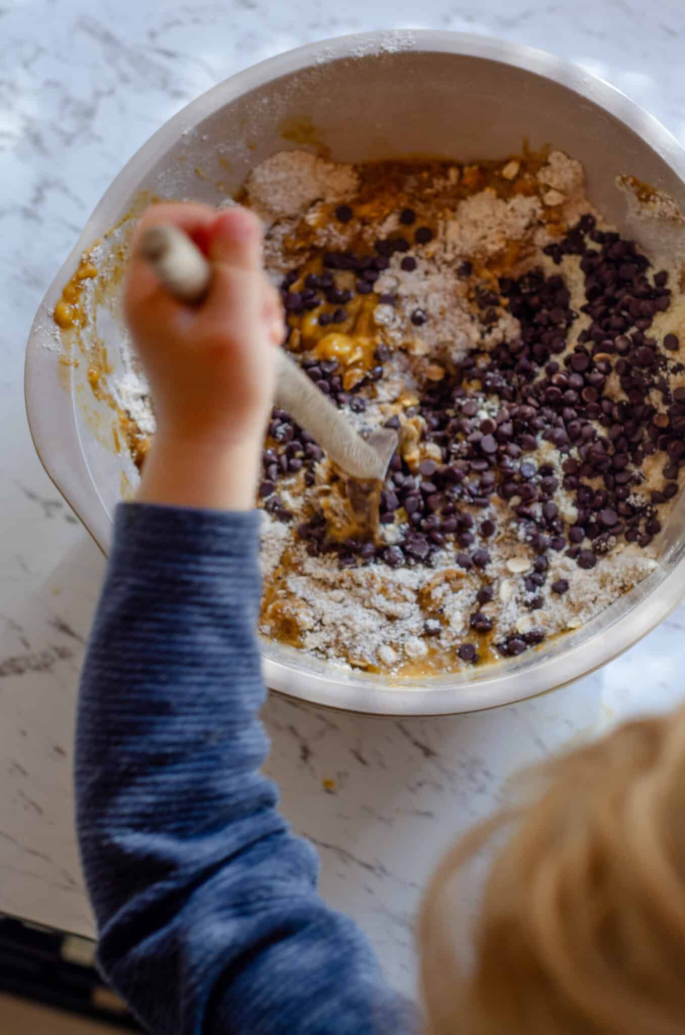 Mixing mini chocolate chips into a flour and banana protein bar with a wooden spoon by the hand of a child.