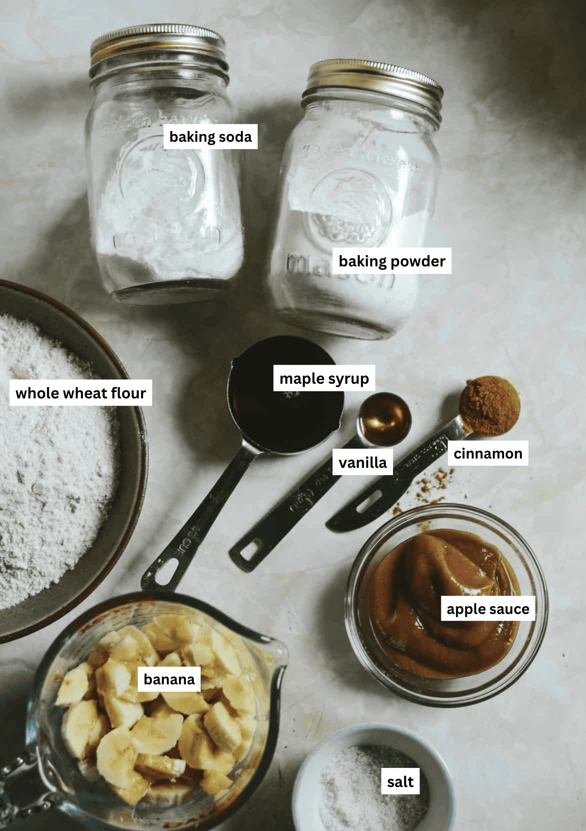 Whole wheat flour, ripe banana. cinnamon, maple syrup, vanilla, applesauce, salt, baking powder, baking soda all on the counter for baking muffins.