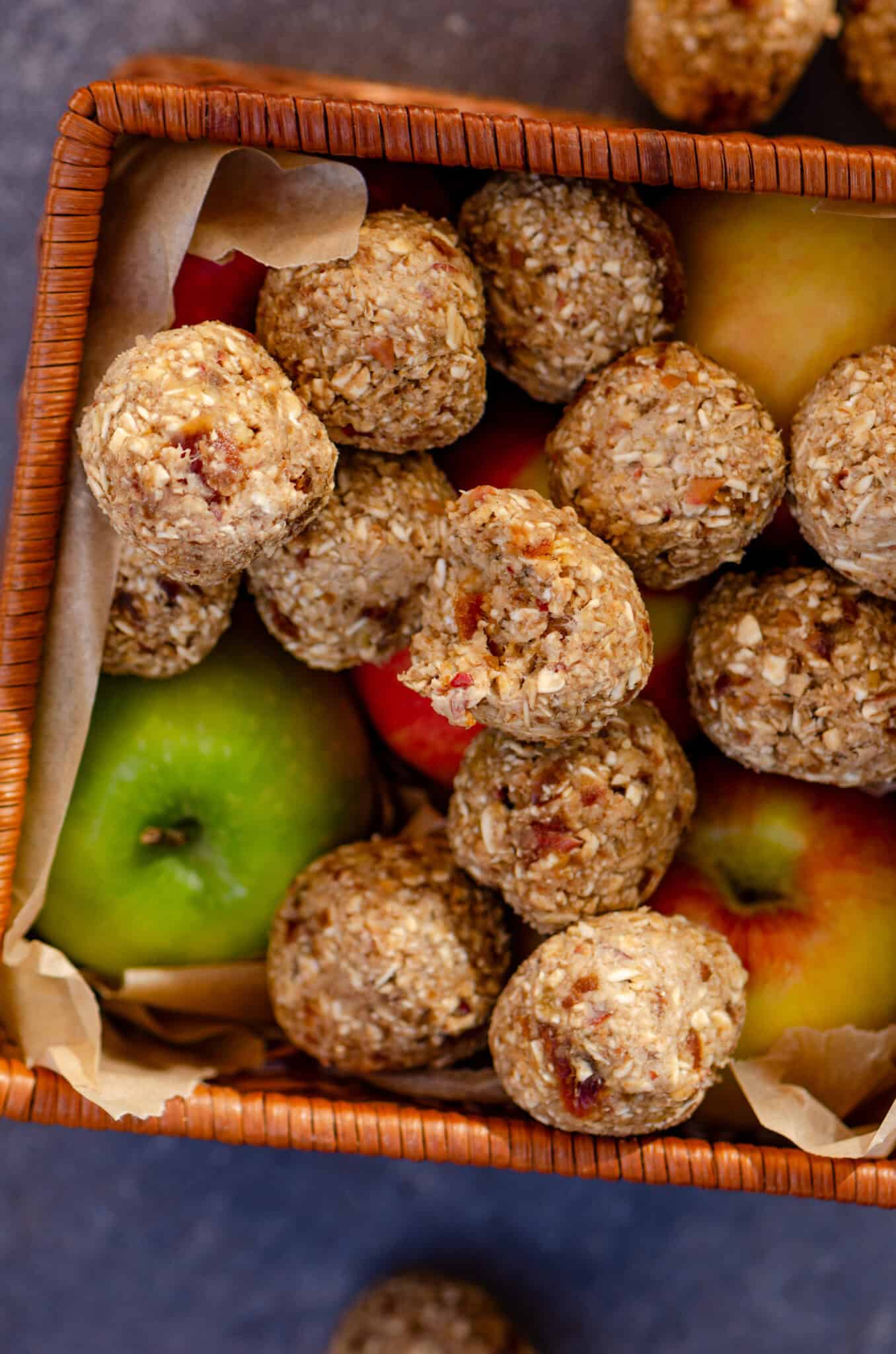 Light brown energy balls with oats and specs of apple in each ball, sitting in a square basket with apples in it.