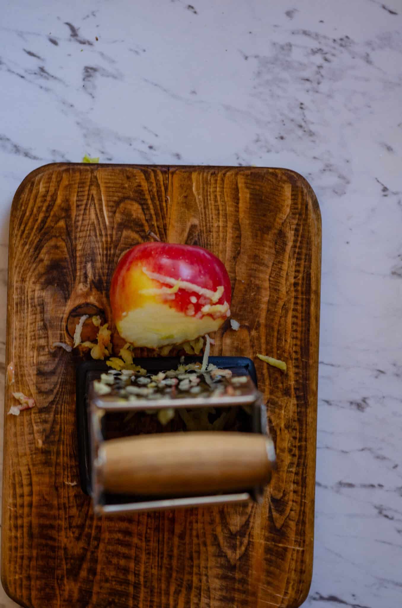Apple being grated on a box grater on top of a small wood cutting board.