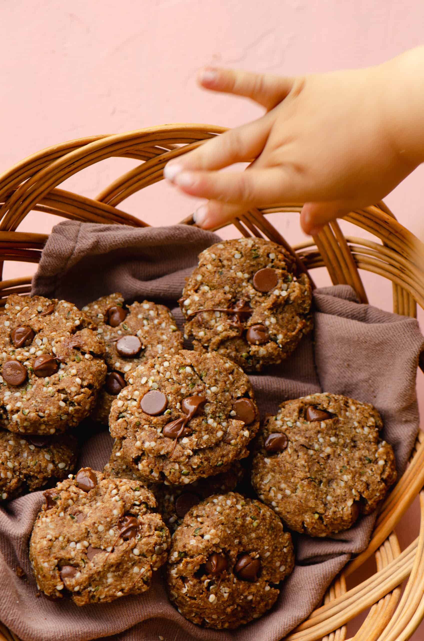 A toddler hand grabbing a brown cookie with hemp hearts and chocolate chips from a basket.
