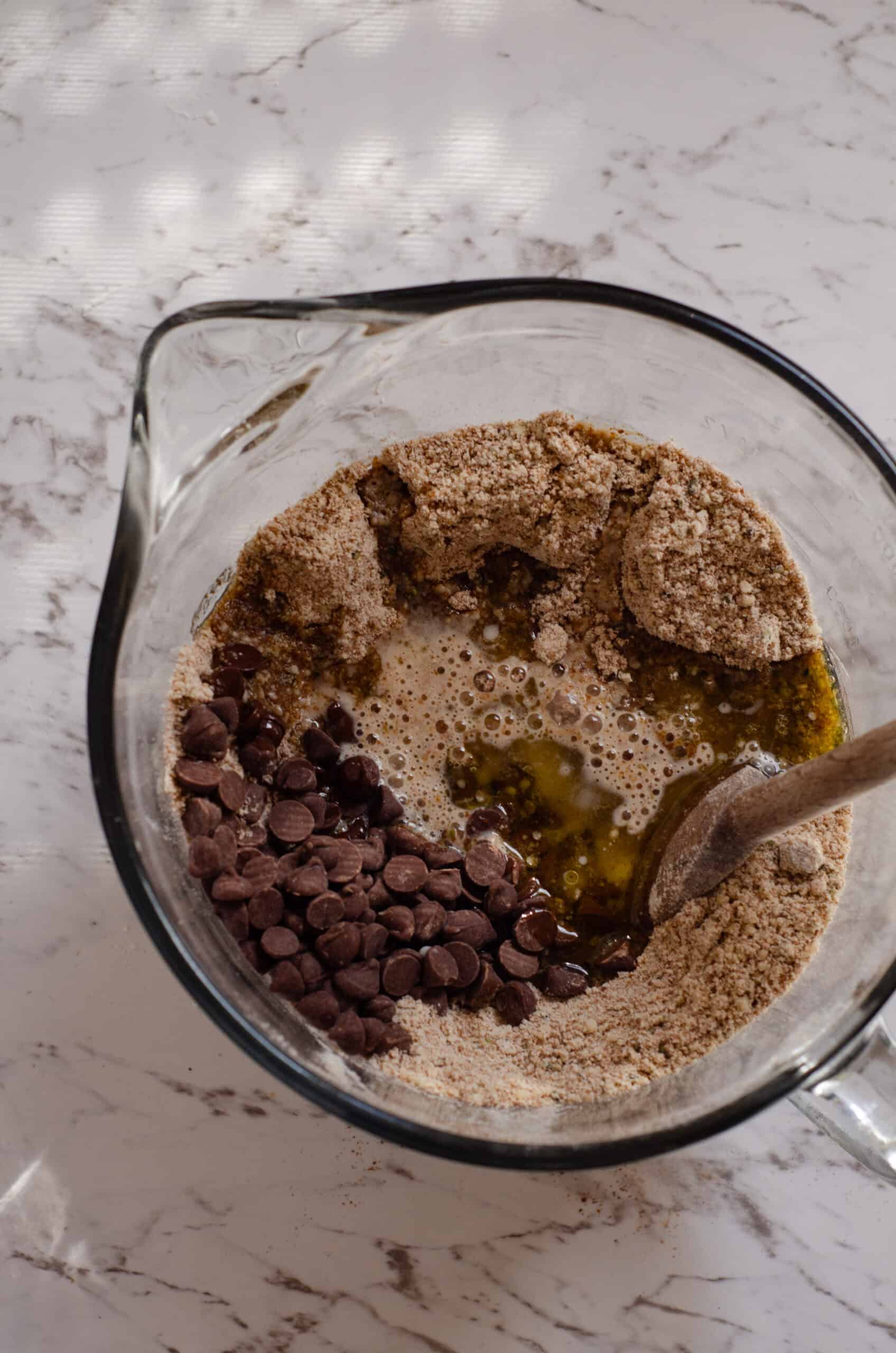 Adding chocolate chips, oil, milk, and egg to bowl of dry ingredients for hemp cookies.
