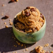 a scoop of light brown cookie dough with chocolate chips in a light blue pottery bowl with a few pieces of cookie dough and chocolate chips on the counter around it.