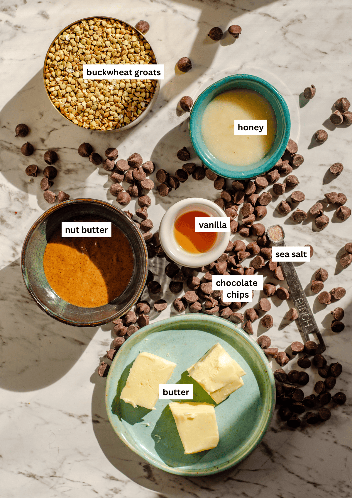 On the counter measured out is honey, nut butter, vanilla, butter, sea salt, and buckwheat, with chocolate chips scattered on the counter.