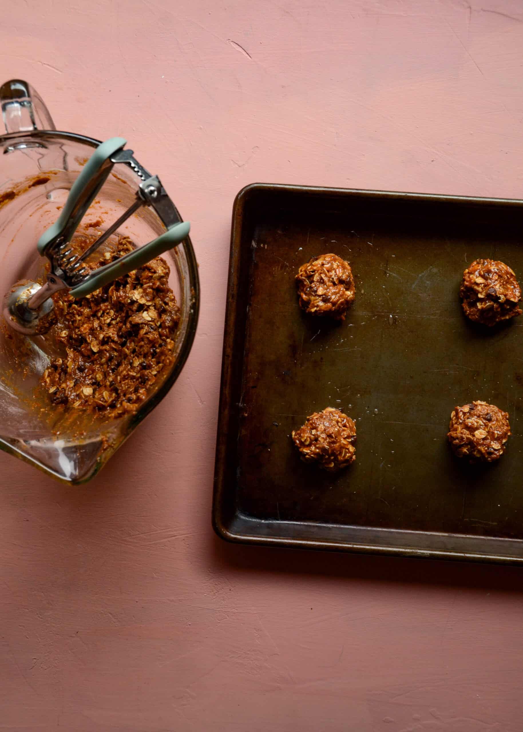 Cookie sheet with balls of cookies dough being scooped from glass mixing bowl with a scoop.