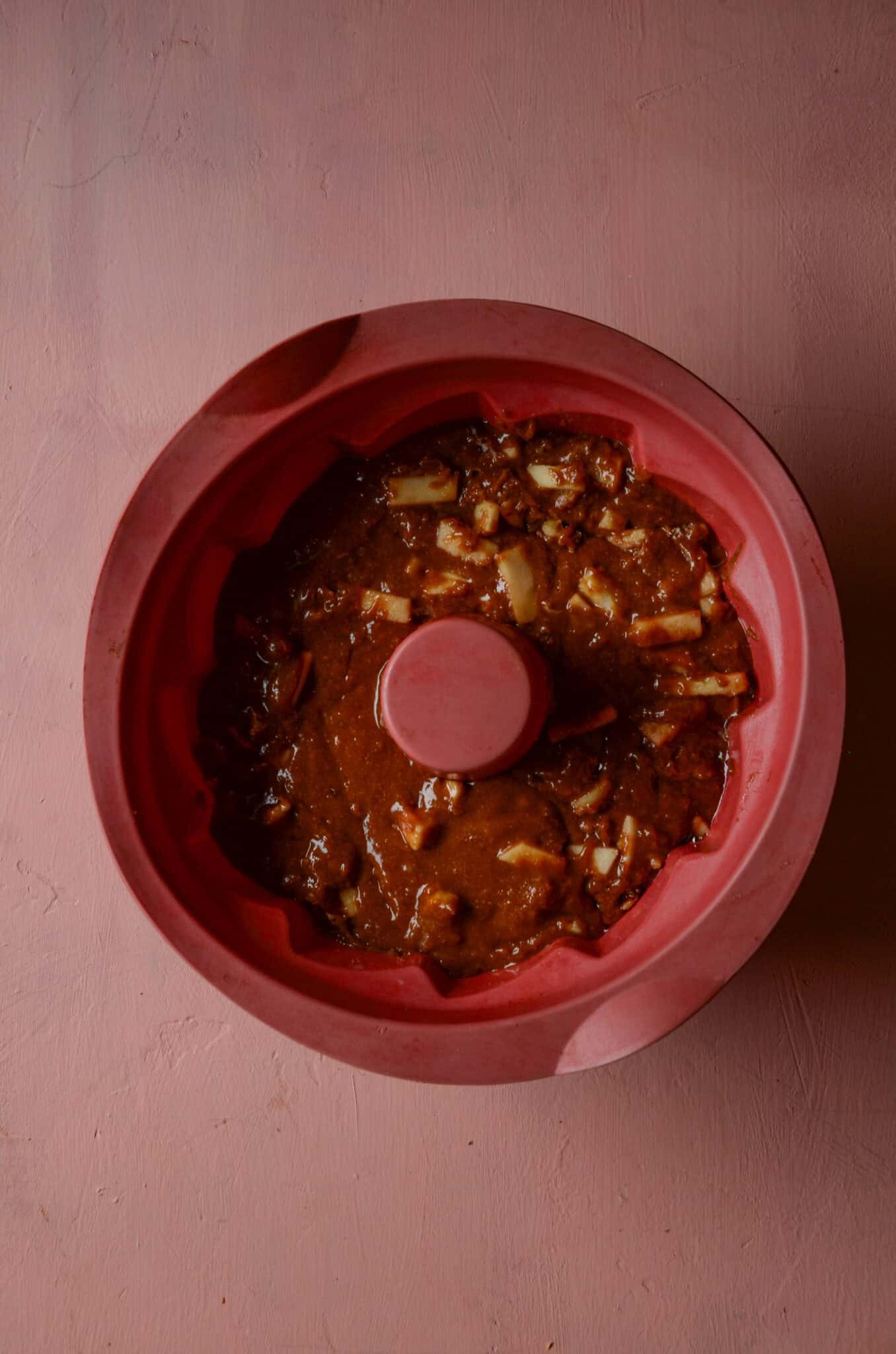 Brown cake batter with white chunks of apples and nuts all in a red bundt pan.
