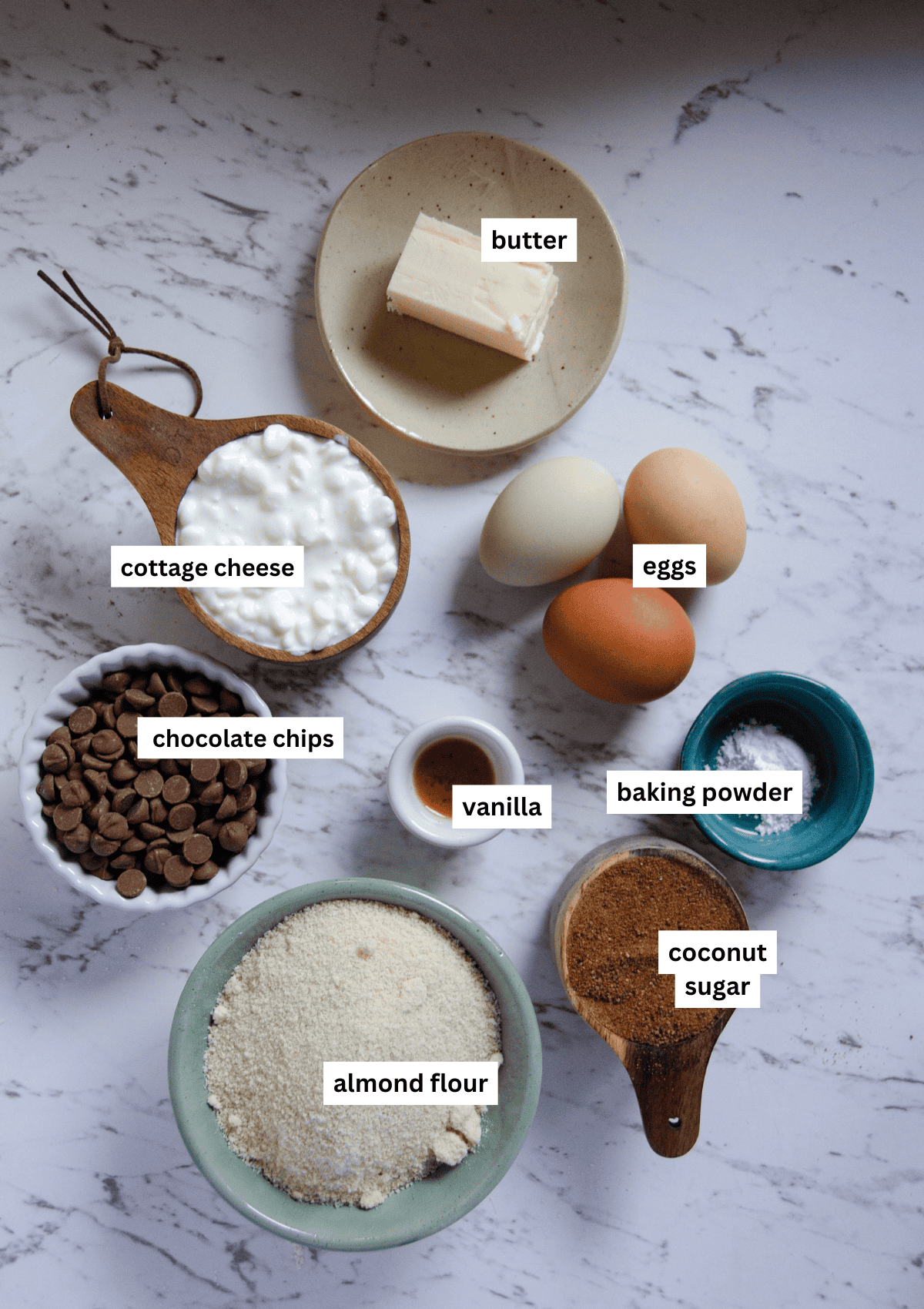 Butter, cottage cheese, chocolate chips, vanilla, almond flour, coconut sugar, baking powder, eggs in bowls.