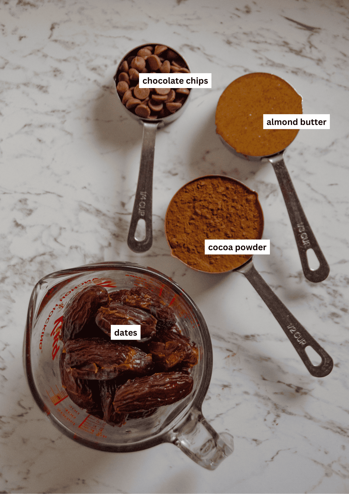A glass measuring cup of dates, and metal measuring scoops with cocoa powder, almond butter, and chocolate chips.