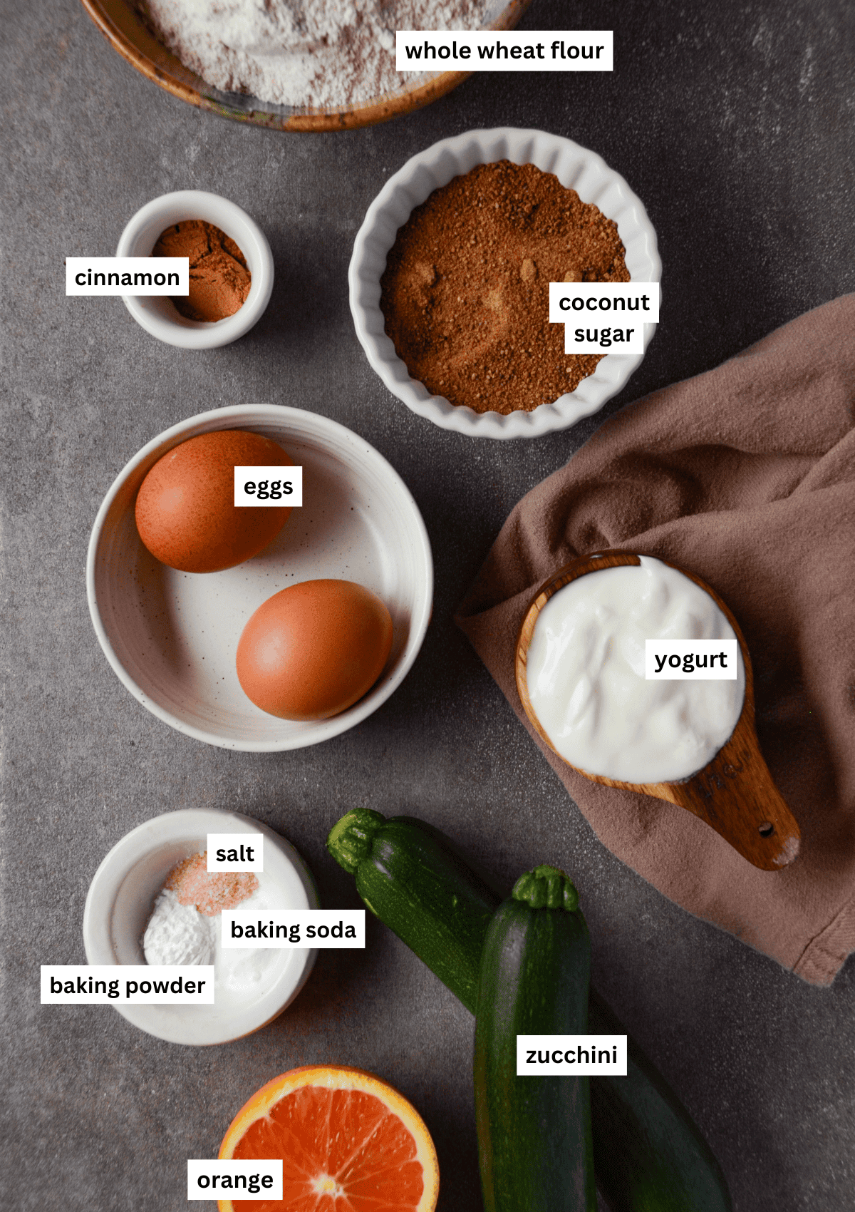 Ingredients in bowls for zucchini muffins including whole wheat flour, vanilla, coconut sugar, eggs, yogurt, sea salt, baking powder, baking soda, zucchini, orange.