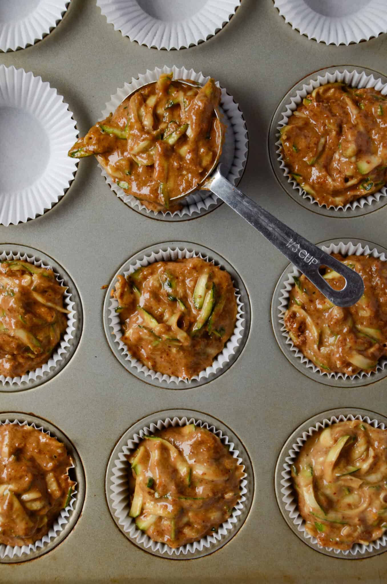 A muffin pan with paper liners with a ⅓ cup measuring scoop putting batter in the pan.