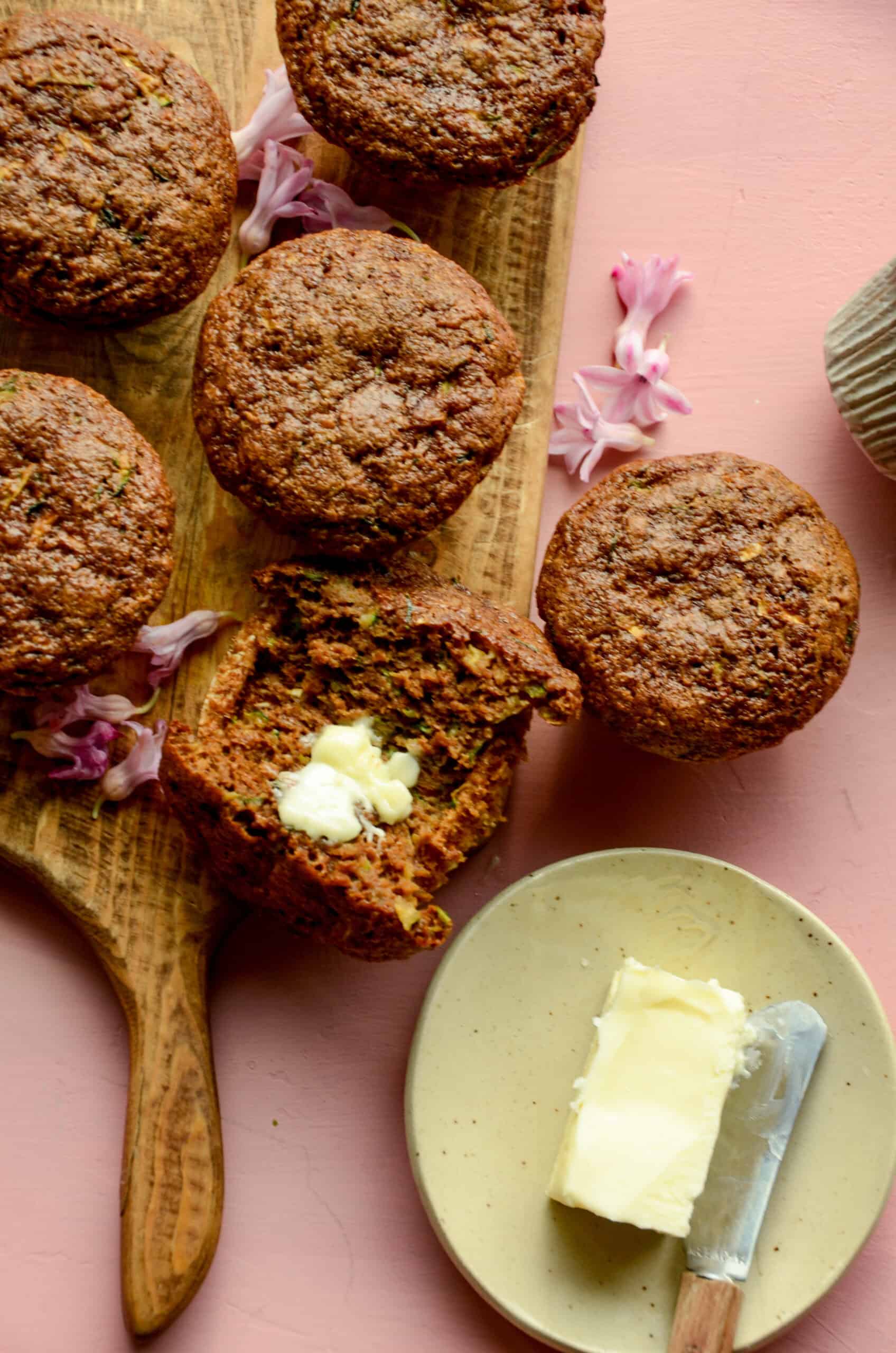 Zucchini muffins on a wooden cutting board, one broken open with a chunk of butter on it.