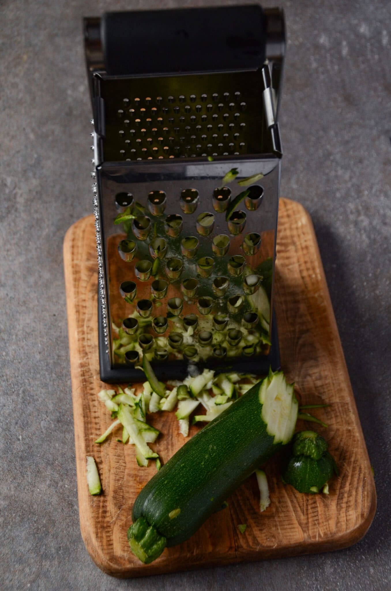 A cheese grater on a cutting board with a zucchini being thinly grated.