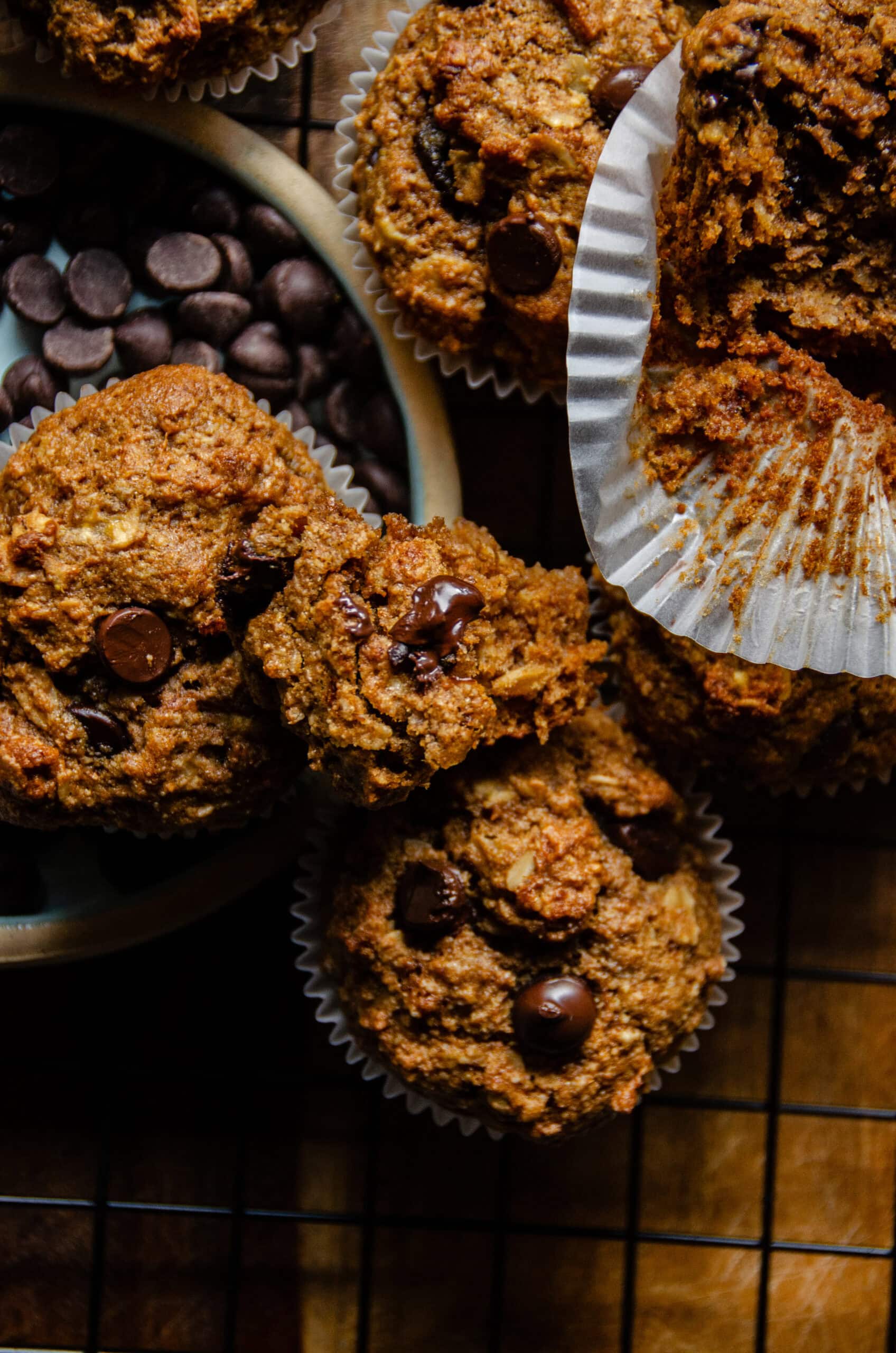 Brown banana muffins in white paper liners, with a chunk of muffin broken off with melted chocolate chips on it.