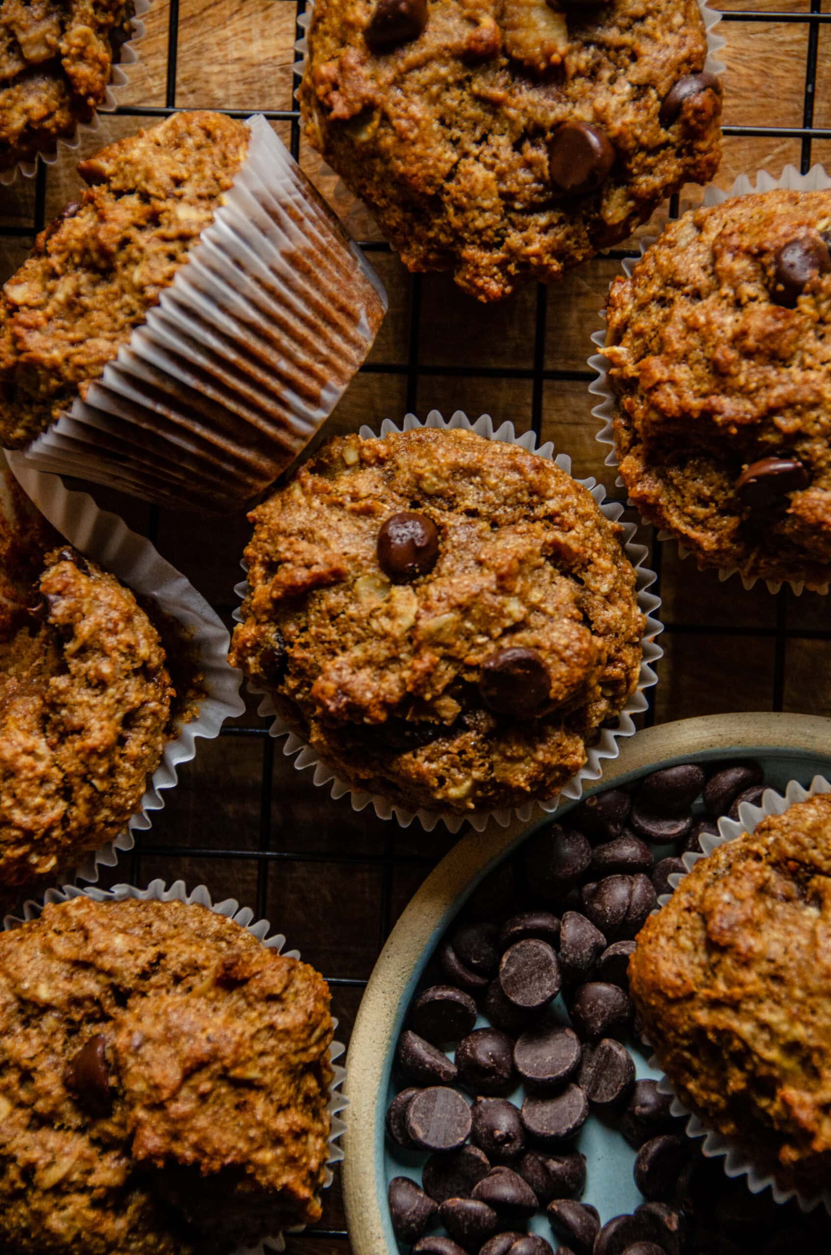 Brown banana protein muffins with chocolate chips sitting on a cooling rack beside a plate of chocolate chip muffins.