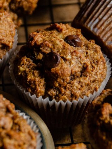 Brown banana protein muffins with chocolate chips sitting on a cooling rack beside a plate of chocolate chip muffins.