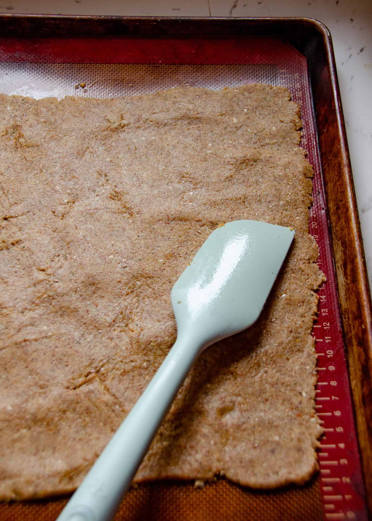 Thick cracker dough spread out of a silicone baking mat on a cookie sheet with a spatula.