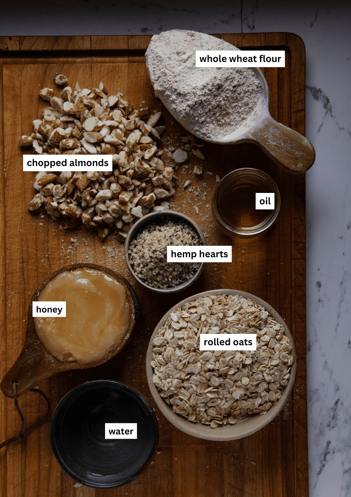 Labelled ingredients in measuring cups and bowls including almonds, whole wheat flour, oil, hemp hearts, honey, rolled oils, water.