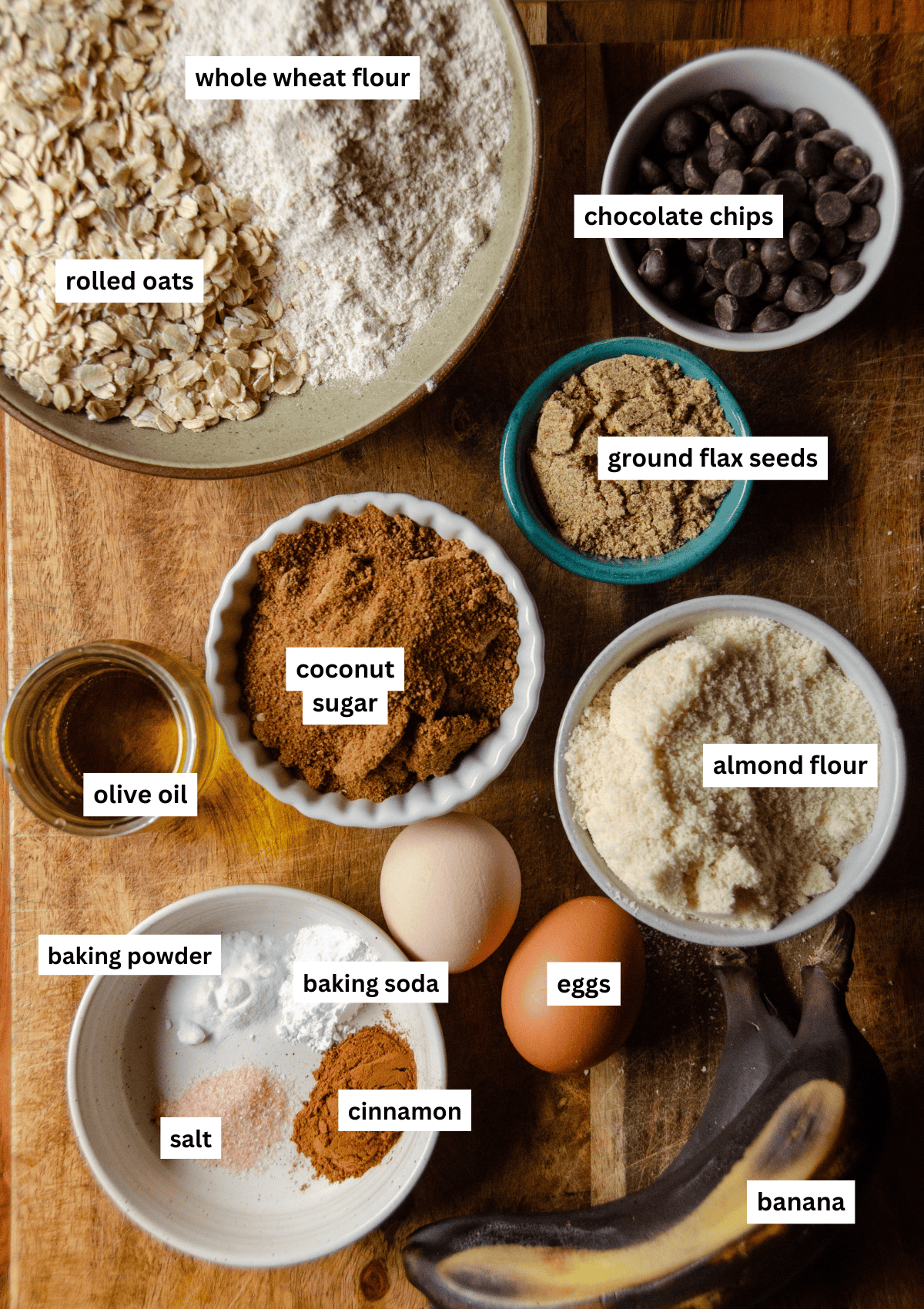 Labeled ingredients for protein muffins in bowls including rolled oats, whole wheat flour, chocolate chips, flax seeds, coconut sugar, vanilla, almond flour, eggs, baking soda, baking powder, salt, cinnamon, overripe bananas.