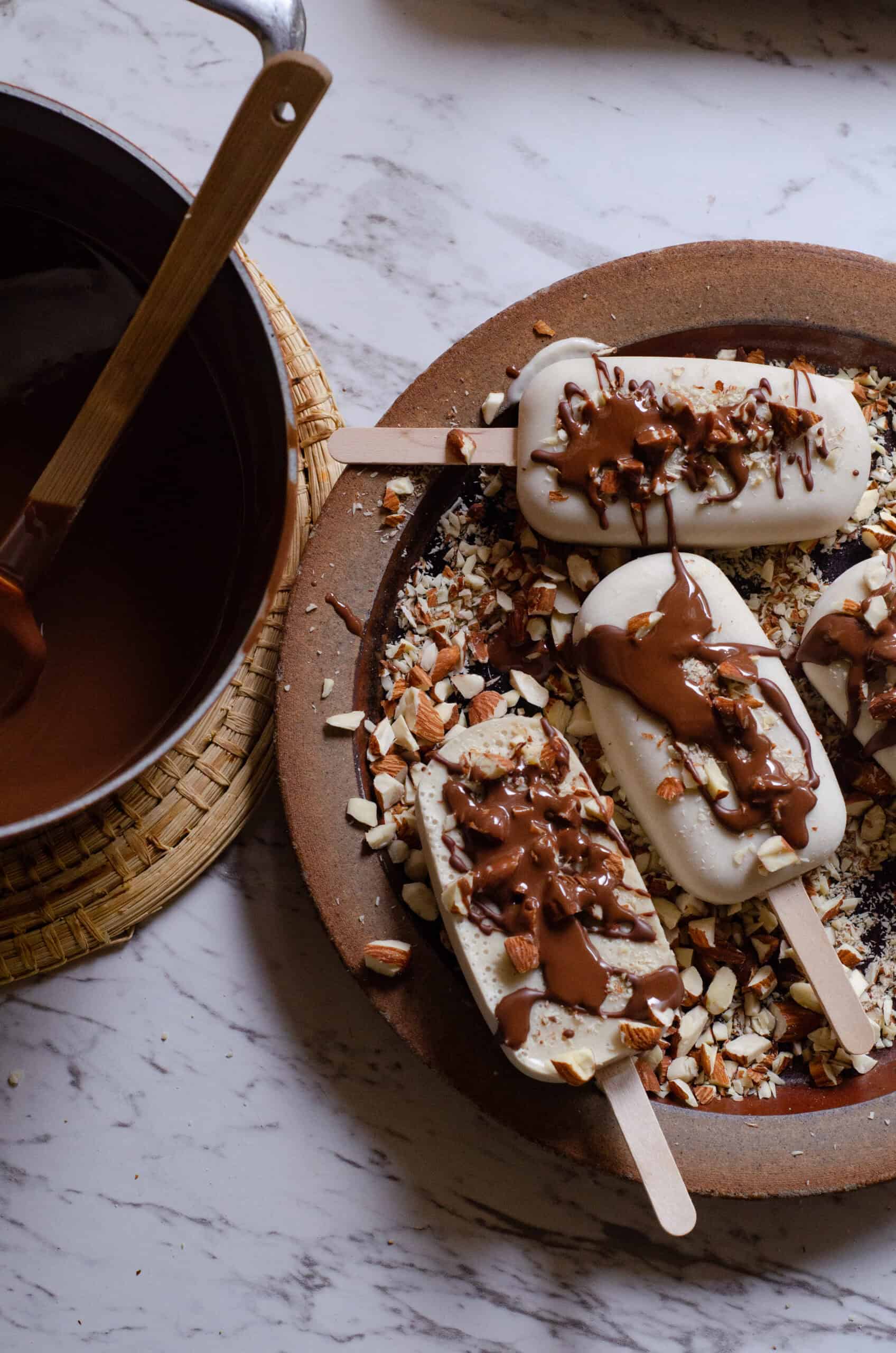 Ice cream bars on a plate with almonds on them and chocolate being drizzled on them.