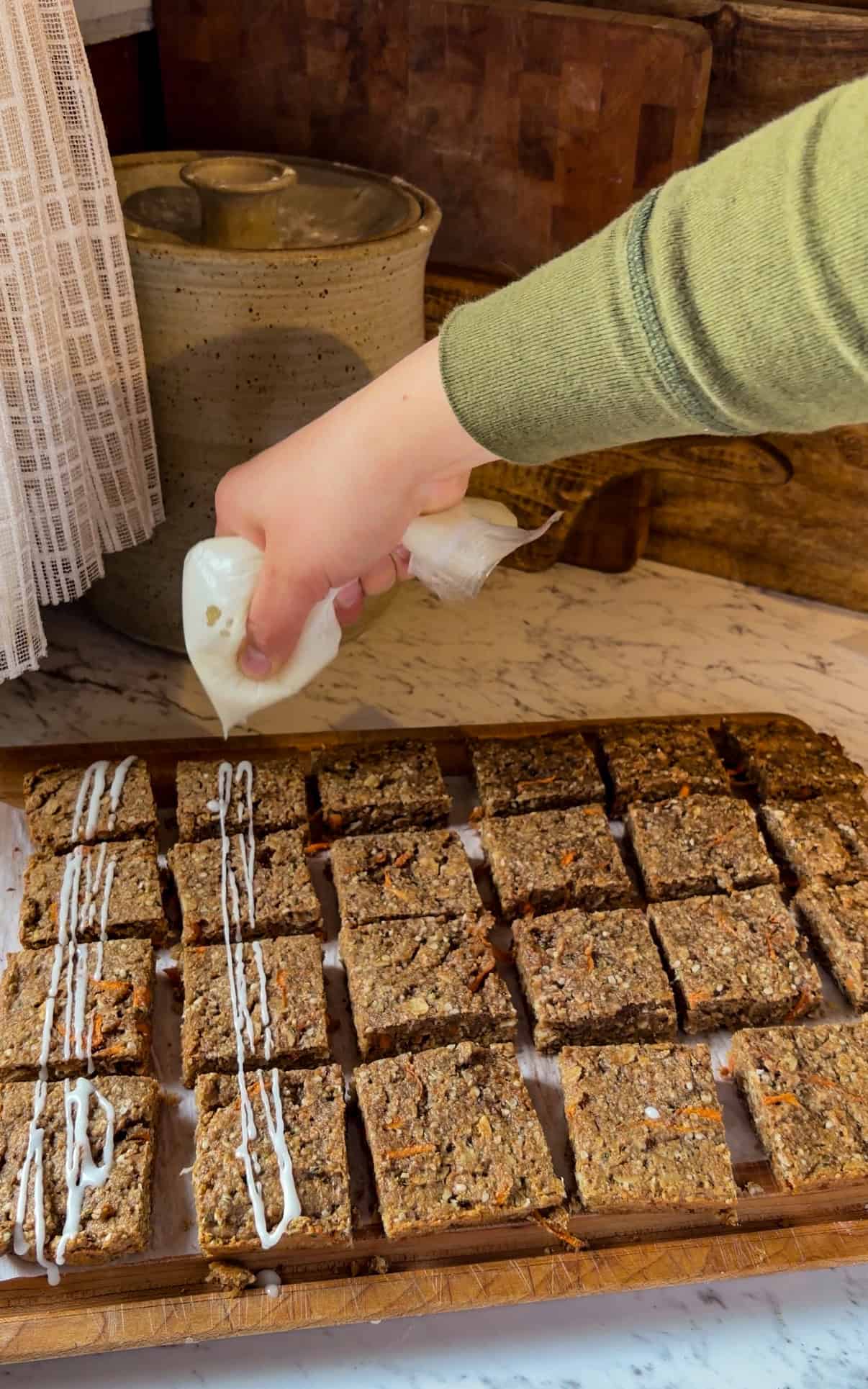 A hand drizzling cream cheese icing on carrot cake protein bars.