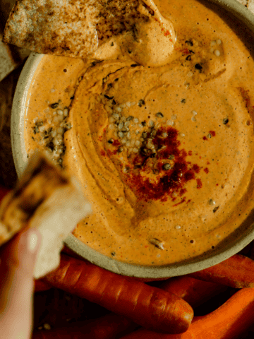 A hand dipping into orange colored protein dip with pitas and carrots beside the bowl.
