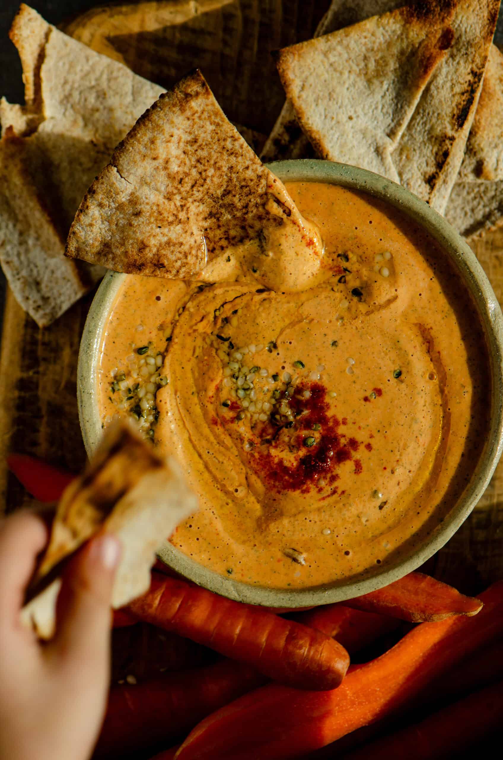 A hand dipping into orange colored protein dip with pitas and carrots beside the bowl. 