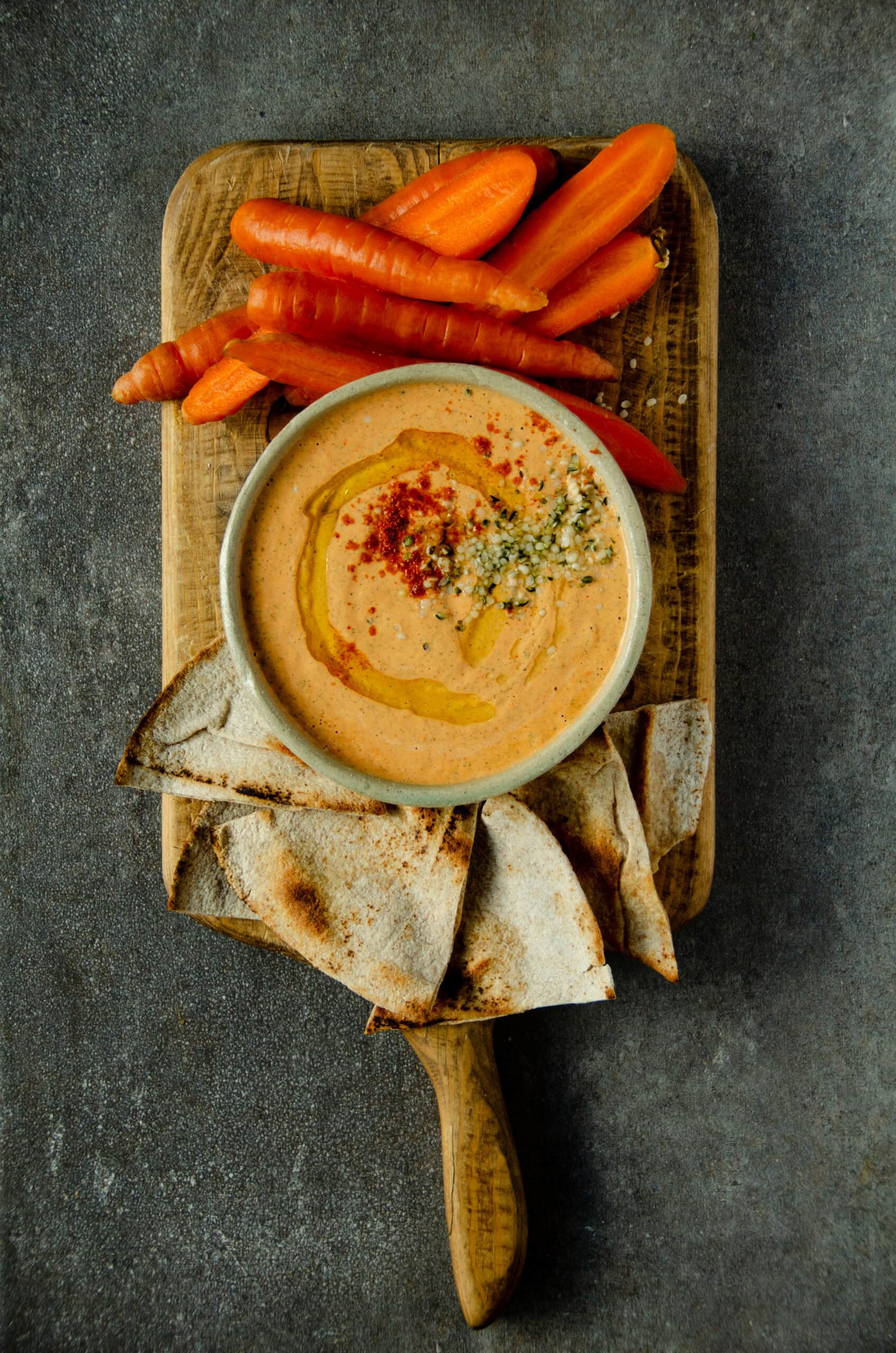 Red pepper protein dip in a bowl on a cutting board with carrots and pita bread.
