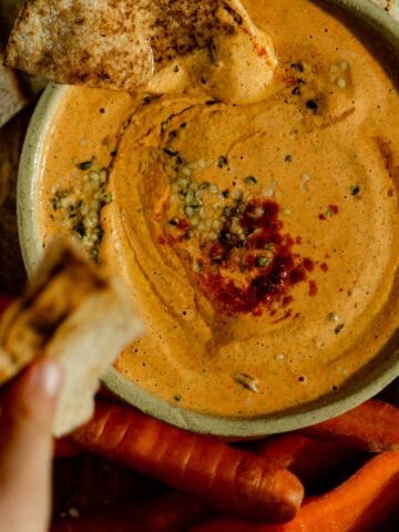 A hand dipping into orange colored protein dip with pitas and carrots beside the bowl.