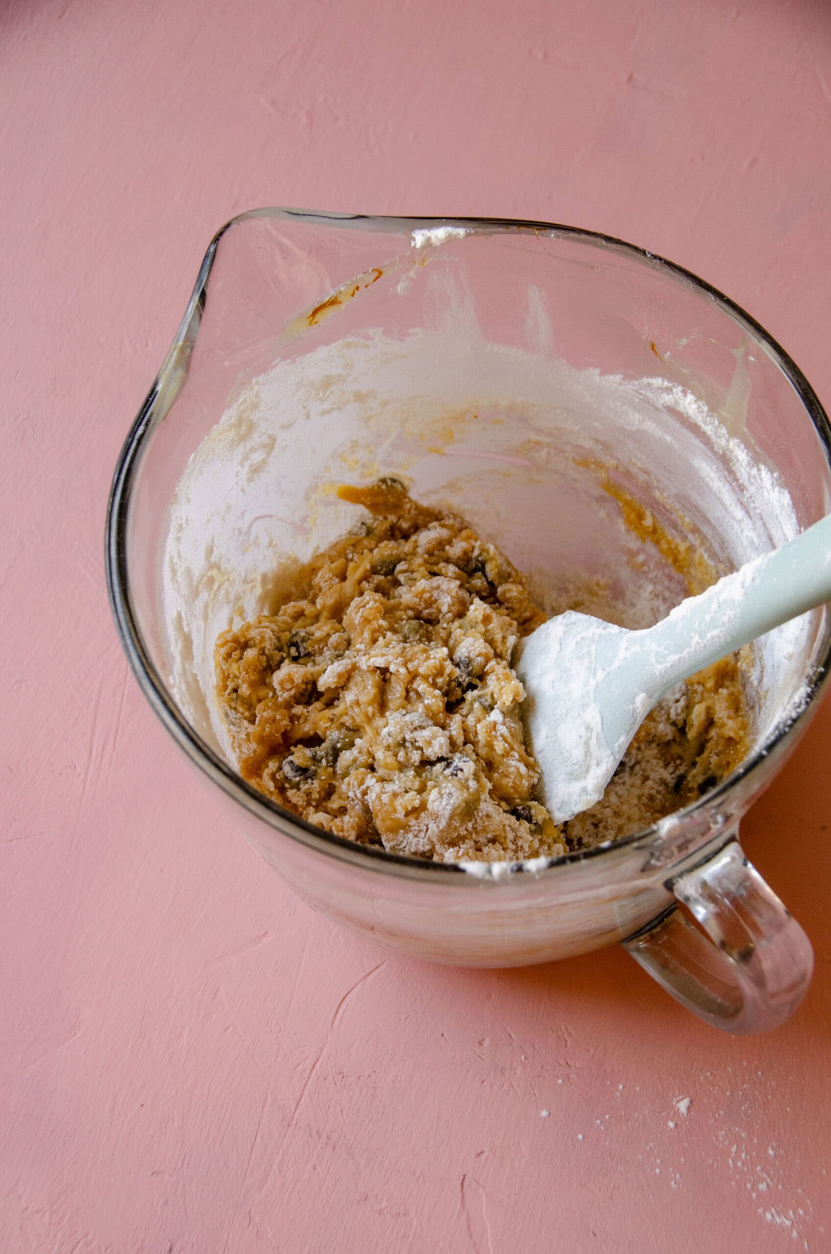 A glass mixing bowl with honey chocolate chip cookie dough and a spatula in the bowl.
