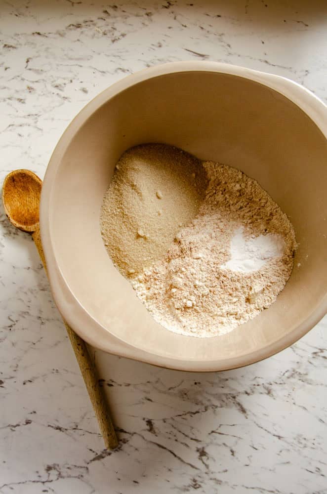 A bowl with sugar and flour, and baking soda and baking powder with a wooden spoon beside it.