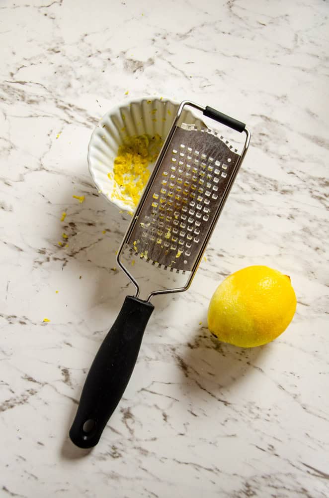 A zester beside a dish with yellow lemon zest in it with a lemon beside it.