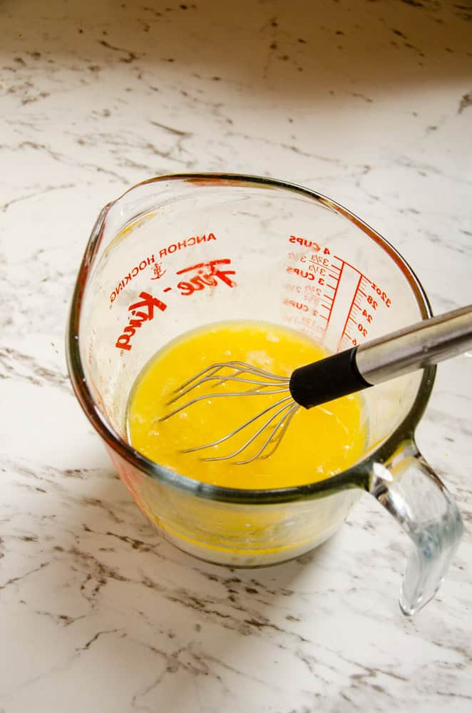 A glass measuring cup with yellow melted butter, and eggs, and a whisk.