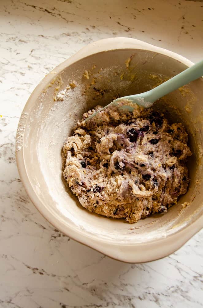 A mixing bowl with wet batter with blueberries in it and a spatula.